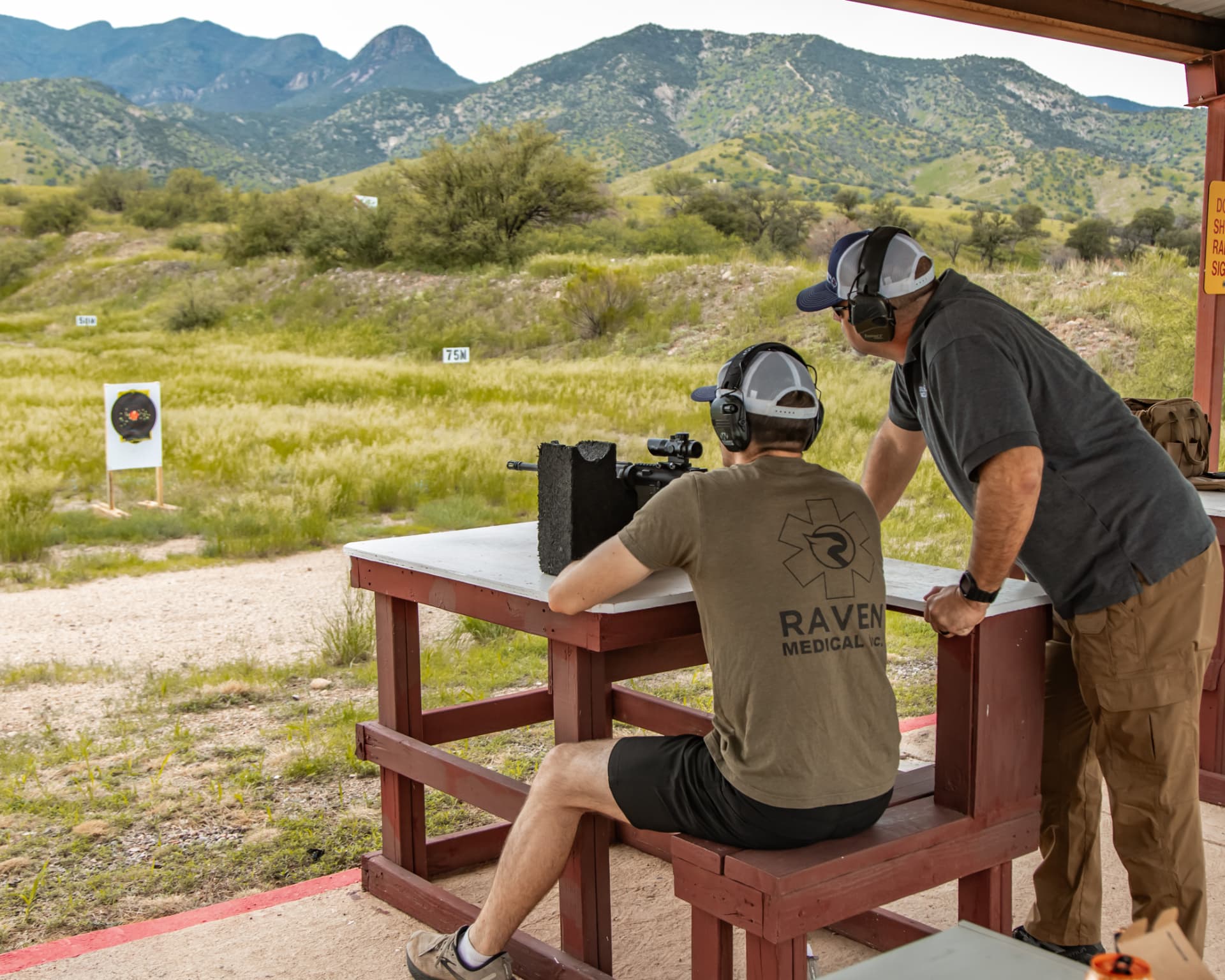 Instructor guiding a student through rifle marksmanship from a bench rest with mountains in the background
